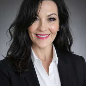 Headshot of Trisha Wright, smiling, with long dark wavy hair, wearing a black blazer and white shirt against a grey background